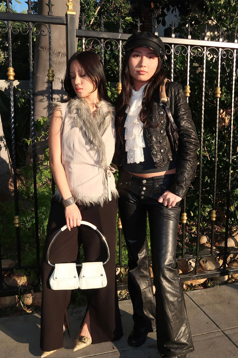 Two women pose in the sunlight against an ornate black iron fence, wearing edgy fall outfits with a fur-trimmed beige top, black flared pants, a leather jacket, and leather trousers. They accessorize with white handbags and statement headwear.