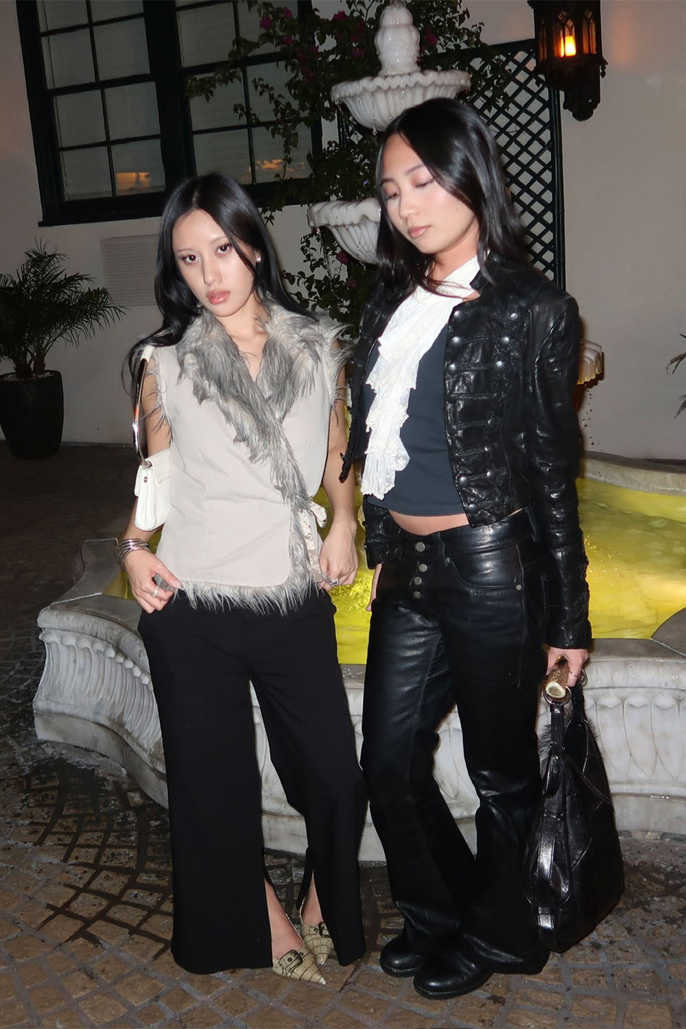 Two women pose beside an illuminated outdoor fountain at night, wearing edgy fall outfits featuring a fur-trimmed beige top, black flared trousers, a black leather jacket, and leather pants.